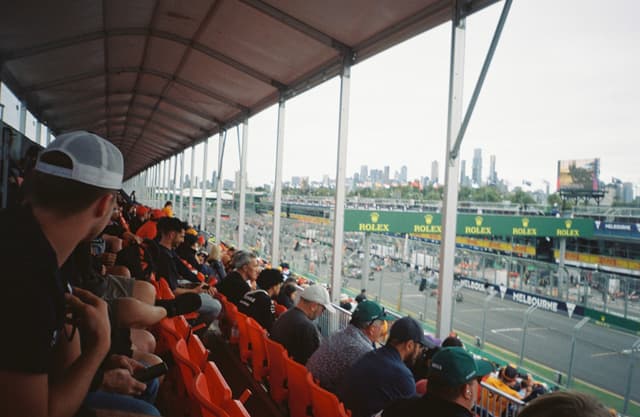 grandstand spectators audience people hats caps seats chairs track racecourse event sports motorsport racing cars vehicles cityscape skyline buildings architecture roof canopy structure barriers fence advertising banners signs green red gray overcast cloudy day outdoor urban environment crowd gathering primary color gray