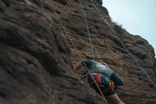 rock climbing climber ropes harness rock wall outdoor adventure sport mountain ascent challenge gear equipment safety helmet nature rugged cliff ascent brown blue sky vertical action physical activity exercise exploration determination focus concentration strength skill technique primary color brown