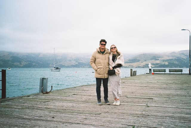 people couple pier water ocean sea boat sailboat mountains hills sky clouds overcast dock railing wood planks jackets coats sunglasses scarf shoes pants jeans beige blue gray white standing outdoors landscape horizon travel vacation leisure calm serene peaceful primary color blue