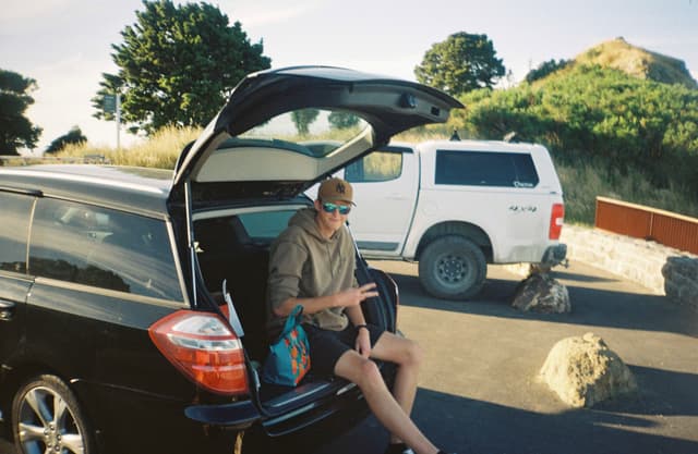 car vehicle person sitting trunk open road parking lot trees sky grass rock hat sunglasses shorts shoes bag black white blue green brown