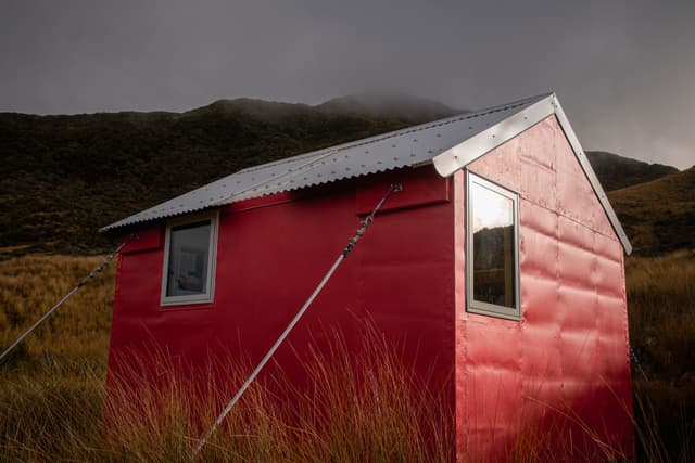 Red hut Cabin Shelter Windows Roof Grass Hillside Mountain Cloudy Sky Outdoors Nature Landscape Remote Rural Simple Rustic Overcast Metal Roof Structure Building Countryside Scenery Wilderness Solitude Tranquil Peaceful Quiet Calm