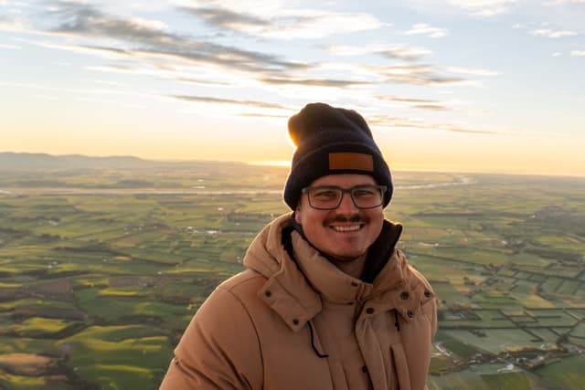 man glasses beanie hat jacket smile landscape field sky clouds sunset sunrise horizon outdoor nature winter warm brown green blue