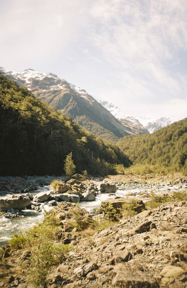 A scenic landscape featuring a river flowing through a rocky valley, surrounded by forested hills and snow-capped mountains under a partly cloudy sky