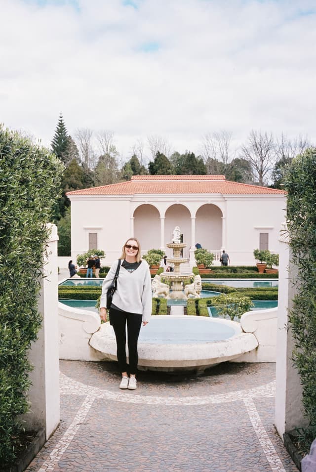 person garden fountain building arch trees sky plants pathway sunglasses sweater pants shoes bag white green blue red stone water architecture bushes walkway