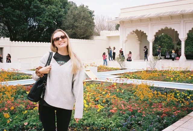 woman sunglasses sweater garden flowers building arches trees people path white green yellow black bag smile outdoors architecture