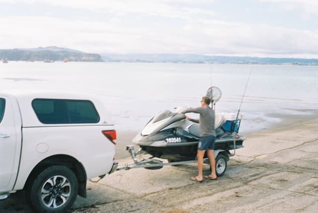 truck trailer jet ski water ocean beach shore fishing rods person sky clouds white blue sand vehicle transport recreation outdoor coast