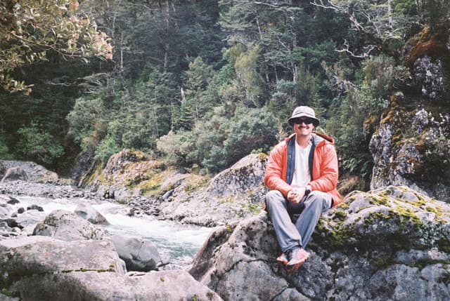 person hat sunglasses jacket orange pants shoes rocks river forest trees greenery nature outdoors sitting primary green