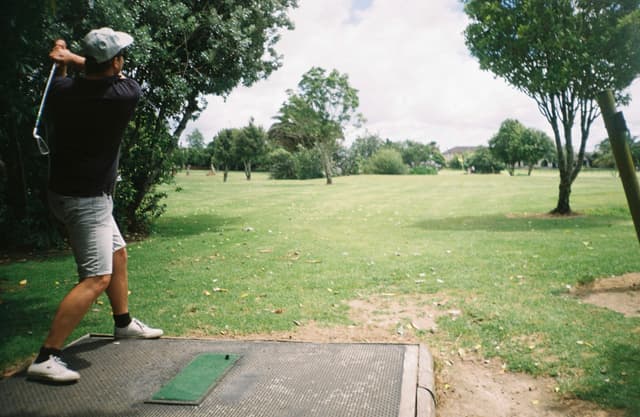 golfer golf swing club course grass trees sky outdoors sports green shorts shoes cap tee box summer fairway player action landscape recreation game activity nature leisure clouds