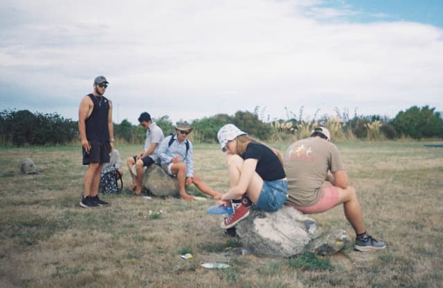 people outdoors grass sky rocks sitting standing hats shoes shorts t-shirts clouds trees field nature casual gathering group summer leisure activity landscape blue green brown gray white relaxed conversation social interaction day light natural environment open space rural scene recreation friendship companionship exploration adventure travel journey relaxation tranquility peacefulness harmony unity connection bonding enjoyment fun freedom simplicity minimalism serenity calmness balance perspective distance horizon openness vastness expanse