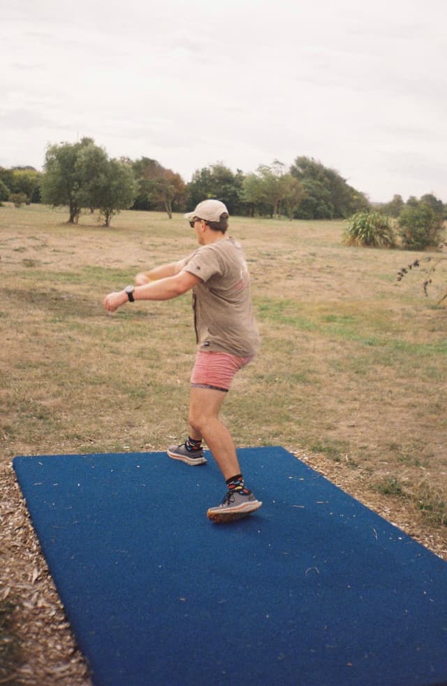 person cap t-shirt shorts shoes watch grass trees sky field outdoors disc golf mat blue brown green standing action sports landscape nature casual summer daytime activity