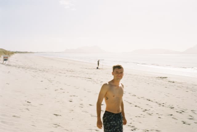 Beach Sand Ocean Water Sky Person Shirtless Shorts Sunlight Horizon Waves Footprints Distant Hills Coastline Shoreline Summer Outdoors Relaxation Leisure Scenic Tranquil Calm Bright Daytime Blue
