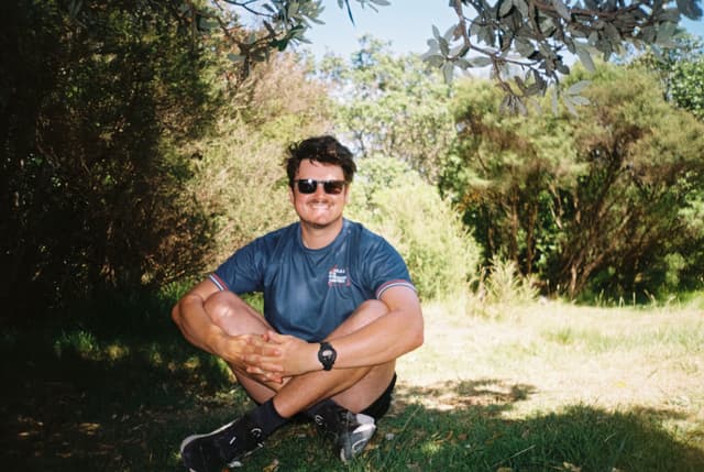 man sunglasses sitting trees grass shorts t-shirt watch shoes greenery outdoors nature sunlight shadow leaves branches summer casual relaxed blue black green brown sunlight forest park garden landscape daytime peaceful serene