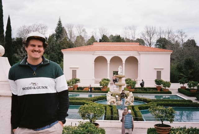 Man Garden Building Fountain Water Trees Bushes Sky Clouds Pathway Columns Archway Hat Jacket Smile Green White Red Stone Grass Shrubs Architecture Outdoors Landscape Sculpture Walkway Balustrade