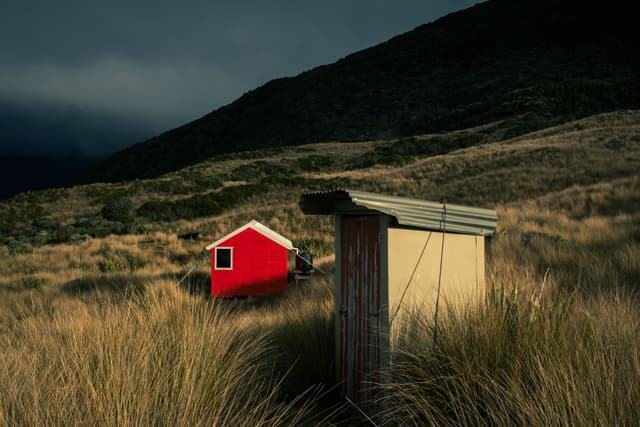 hut cabin grass field mountain hill sky clouds red brown green landscape nature outdoor rural scenery building structure shadow moody dark overcast wilderness remote solitude peaceful tranquil serene primary color
