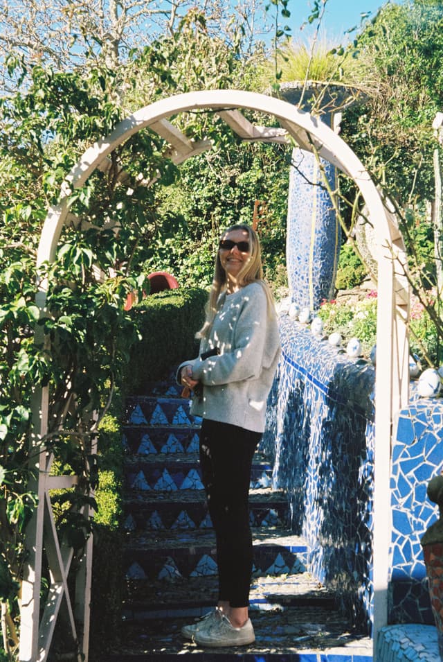 arch garden plants woman sunglasses pathway stairs blue tiles mosaic greenery sunlight outdoor casual clothing sneakers primary blue