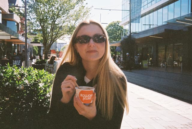 woman sunglasses ice cream spoon cup outdoor street building tree plants sunlight shadow casual clothing blonde hair long hair urban scene daytime primary color green