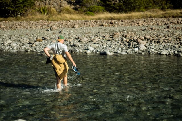water river rocks person backpack hat shoes outdoors nature walking stream blue green brown sunlight trees grass pebbles shore wading landscape adventure exploration travel wilderness scenery clear day summer casual clothing environment tranquility relaxation leisure activity journey exploration calm peaceful serene natural beauty rugged terrain exploration spirit journeying exploring crossing ford ford crossing