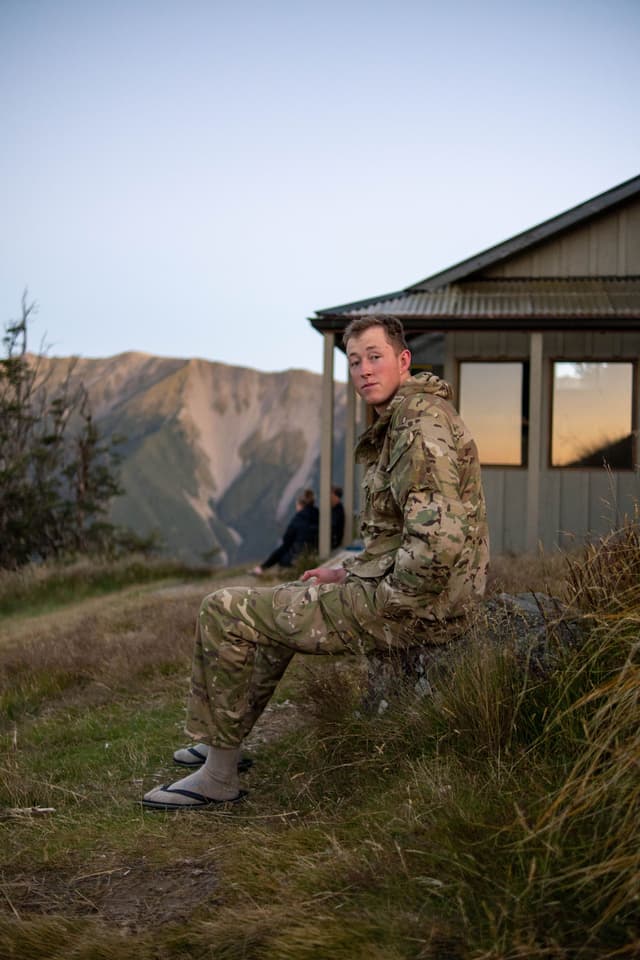 person camouflage clothing house grass mountain sky outdoor sitting evening landscape nature building window brown green blue