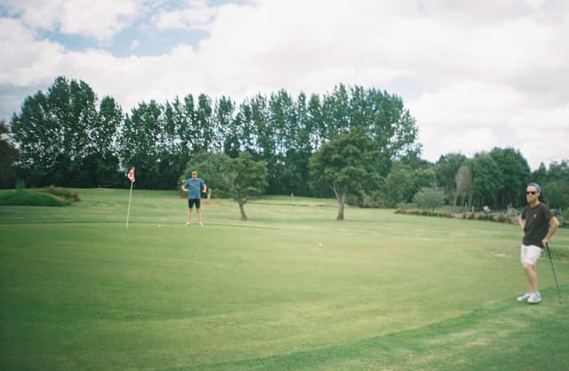 golf course grass trees sky flag pole people outdoors green blue clouds landscape sports leisure nature field fairway golfer putting hole summer day activity recreation scenery