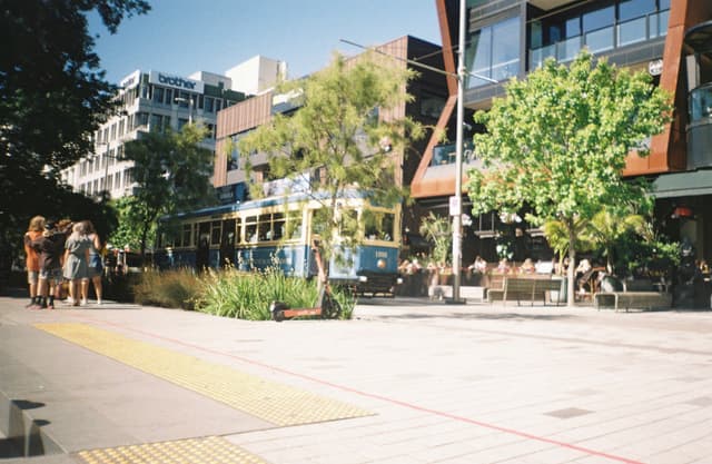 Street Tram Building Trees People Sidewalk Blue Sky Windows Architecture Urban Cityscape Greenery Shadows Sunlight Outdoor Transportation Pathway Modern Shops