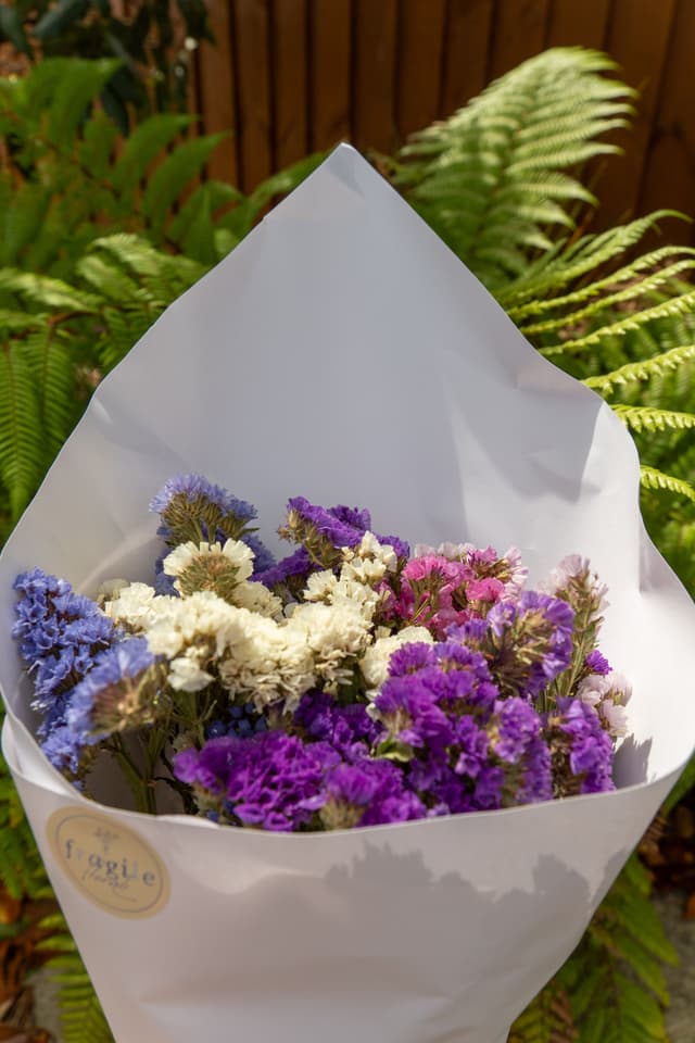 flowers bouquet purple white green leaves ferns paper wrap sunlight garden nature wooden fence outdoors floral arrangement blossoms petals stems natural beauty vibrant fresh decorative gift present colorful bright elegant delicate lush foliage background serene peaceful romantic primary color