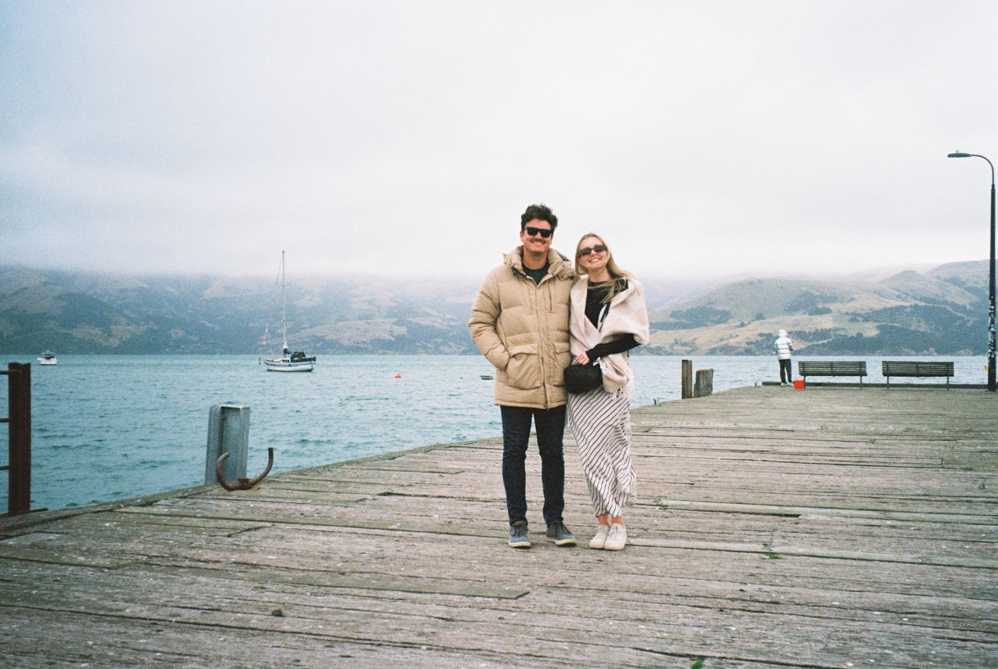 people couple pier water ocean sea boat sailboat mountains hills sky clouds overcast dock railing wood planks jackets coats sunglasses scarf shoes pants jeans beige blue gray white standing outdoors landscape horizon travel vacation leisure calm serene peaceful primary color blue