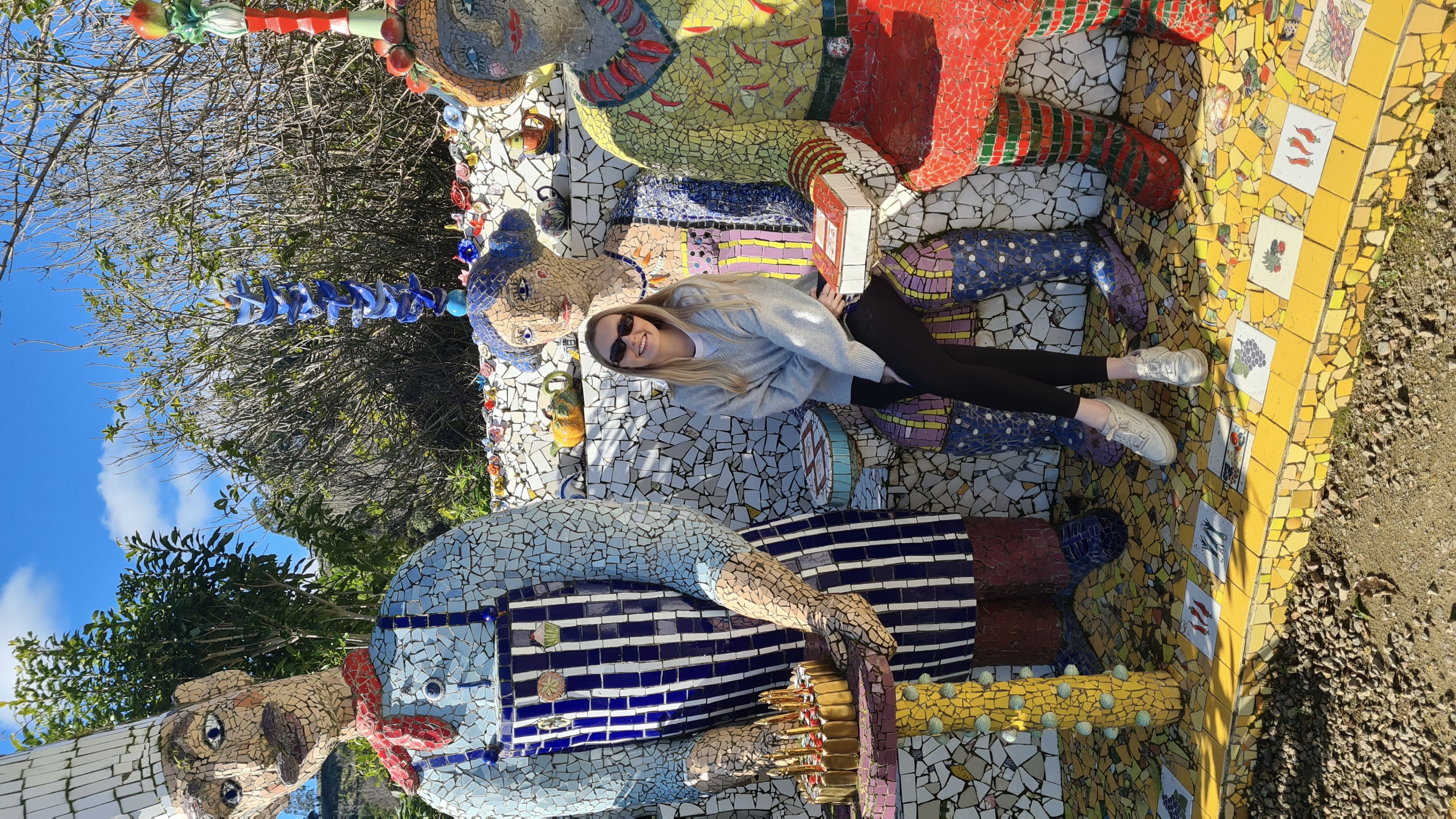 sculpture mosaic art people figures colorful blue sky trees outdoor bench sitting sunglasses hat apron red yellow green tiles stone