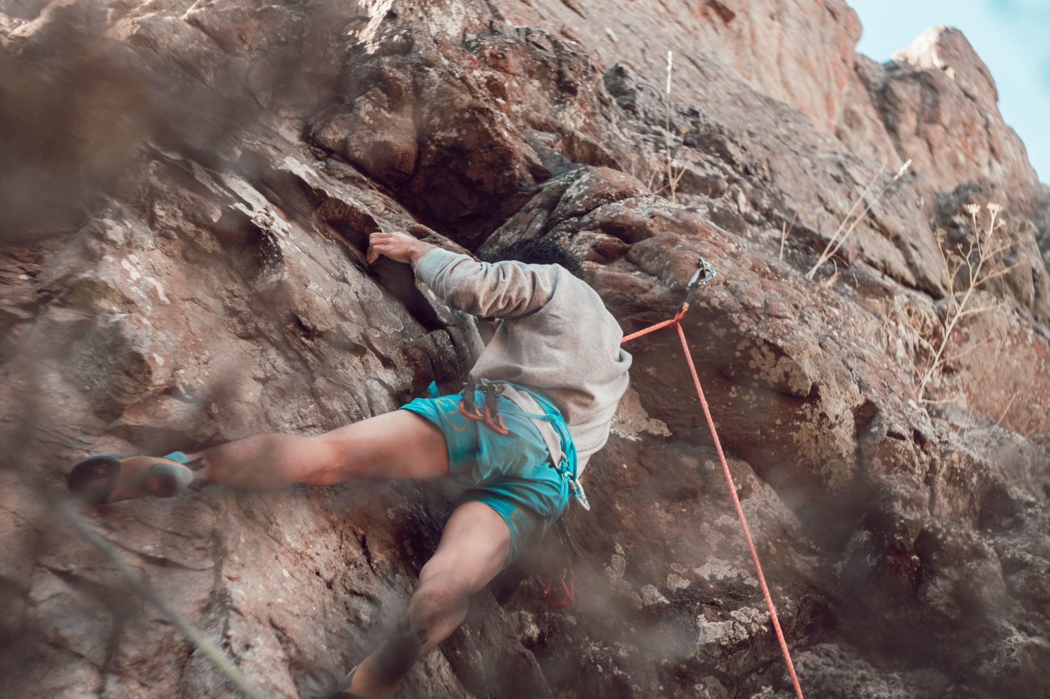 rock climbing climber rope harness rock cliff outdoor adventure sport safety helmet shorts shoes sky blue brown rugged ascent challenge vertical nature equipment action movement effort concentration determination exploration fitness strength technique balance grip foothold handhold adrenaline recreation physical activity skill perseverance courage focus landscape terrain climbing gear primary color brown