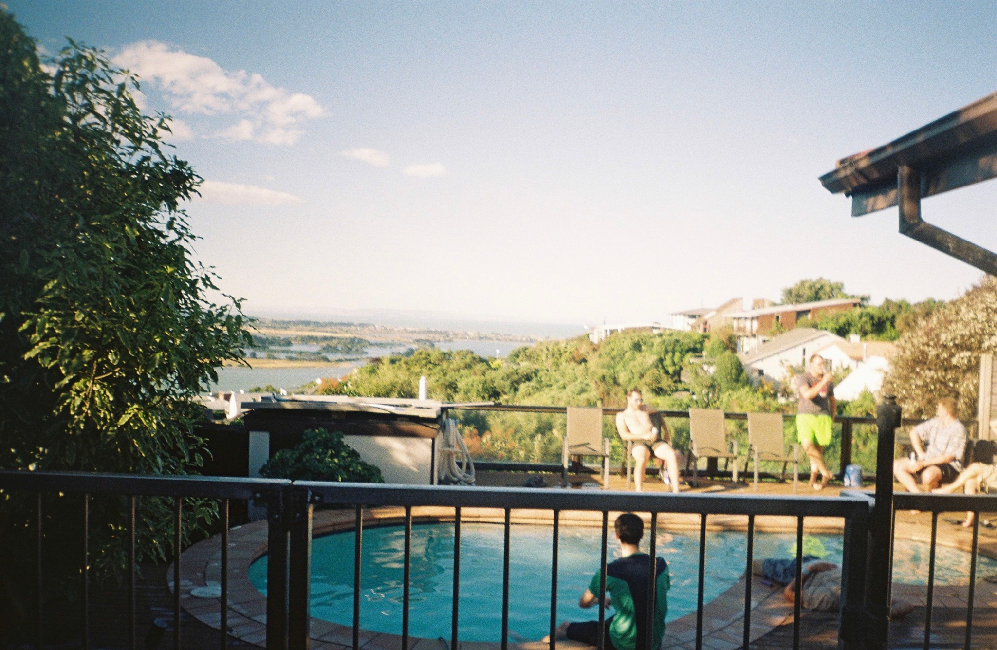 pool fence people trees sky clouds water chairs house railing greenery outdoor blue