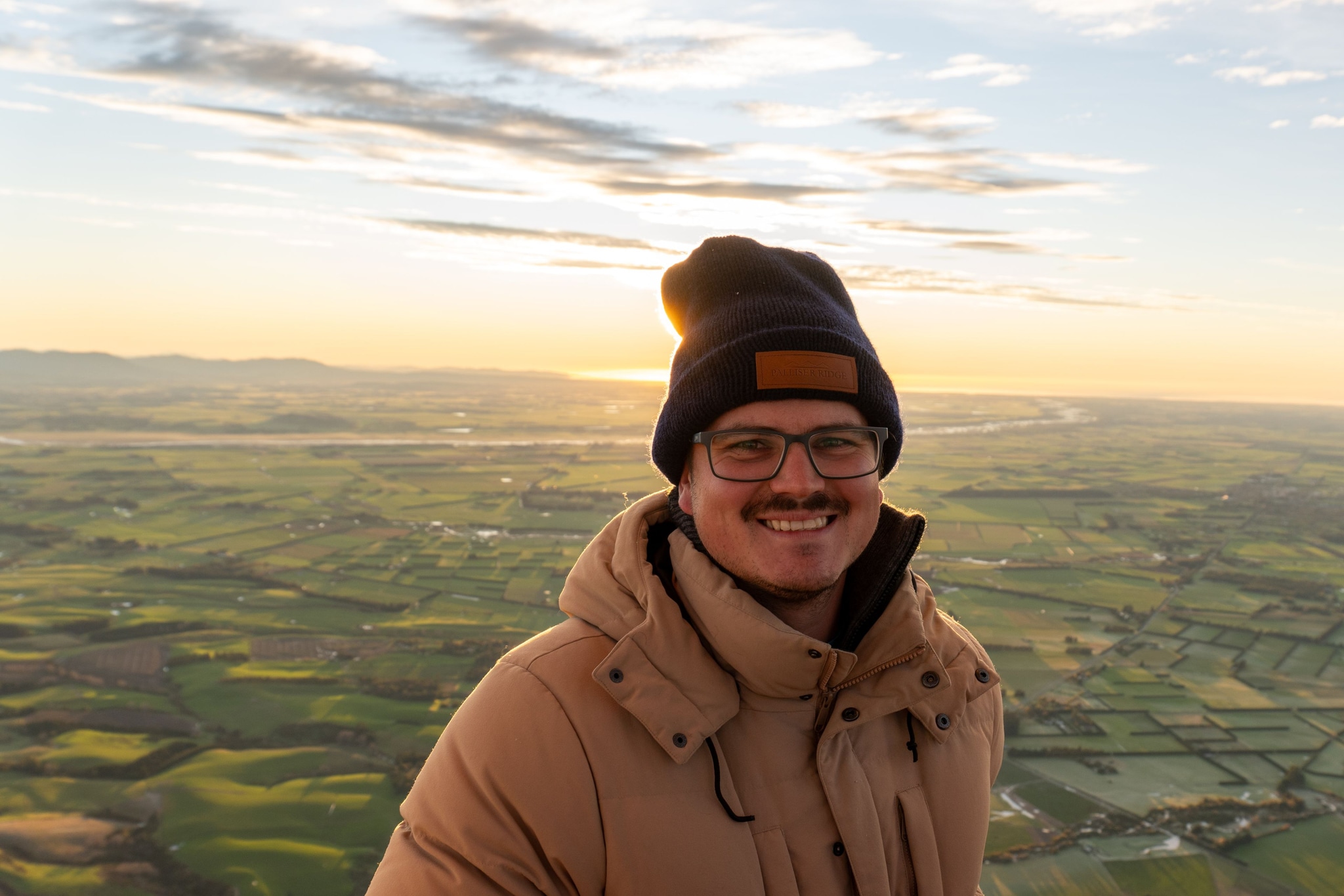 man glasses beanie hat jacket smile landscape field sky clouds sunset sunrise horizon outdoor nature winter warm brown green blue