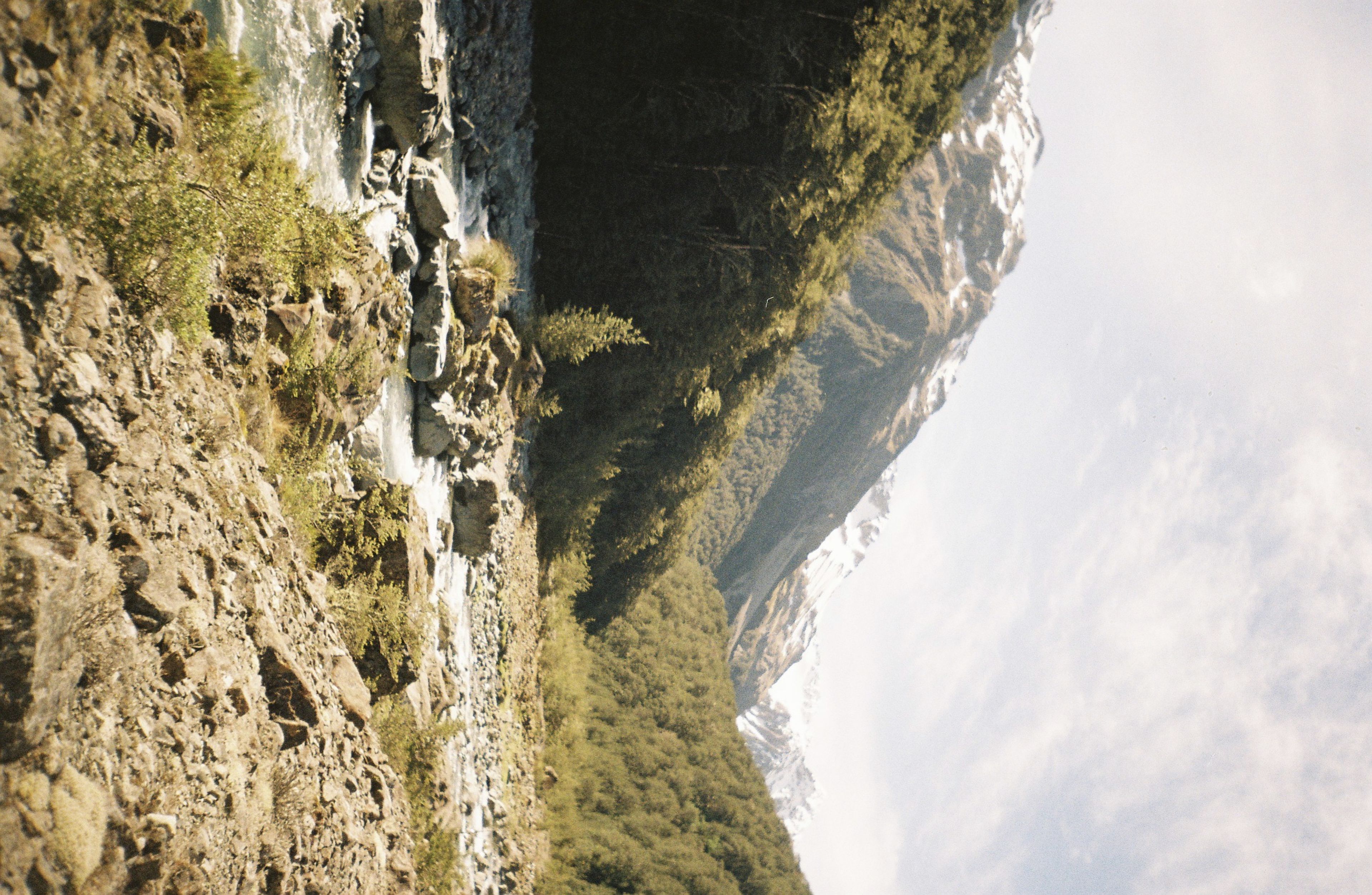 A scenic landscape featuring a river flowing through a rocky valley, surrounded by forested hills and snow-capped mountains under a partly cloudy sky