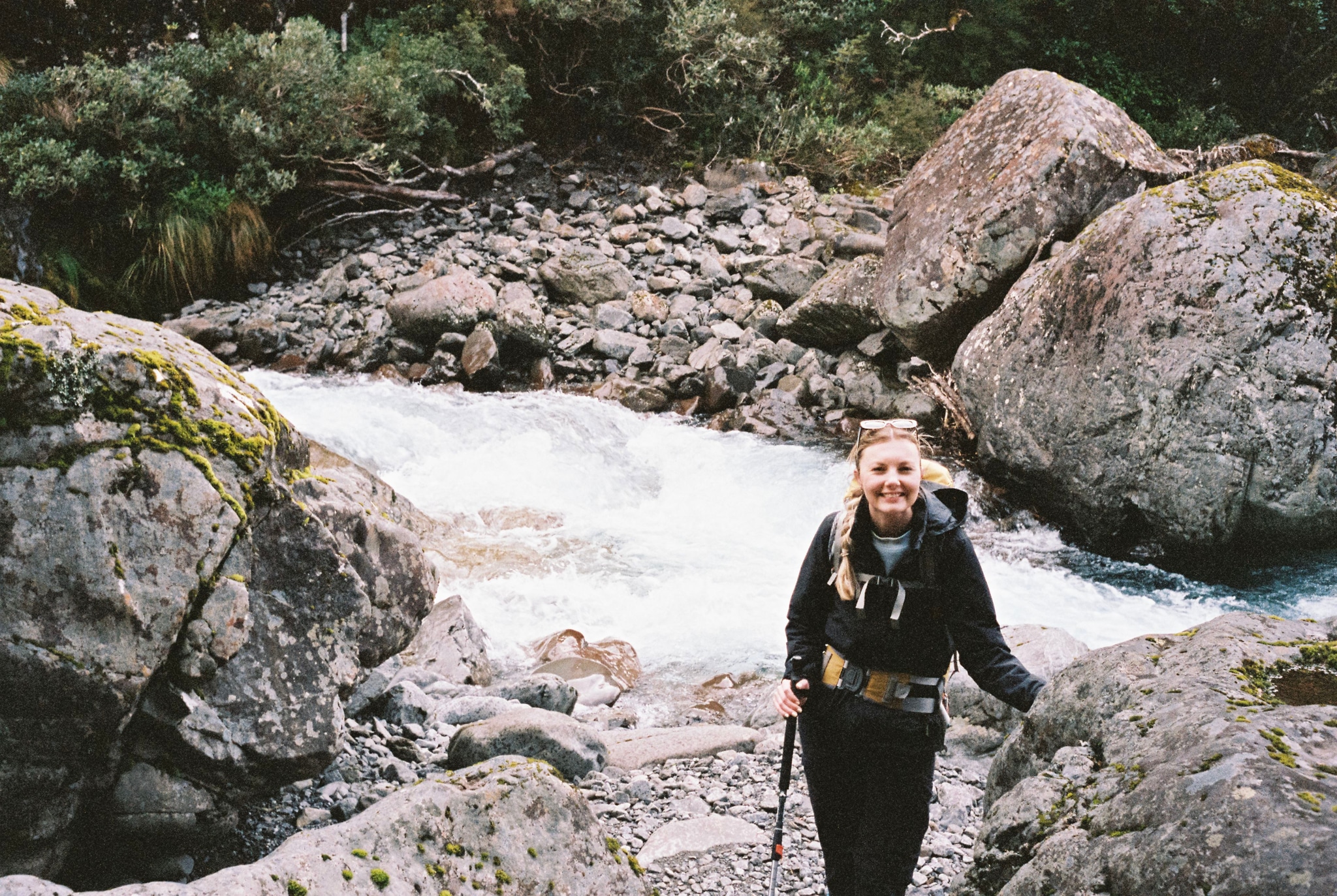 river rocks person hiking backpack nature outdoors forest stream water boulders greenery moss trees landscape adventure trekking exploration wilderness travel scenic trail blue gray white flowing current path journey environment natural beauty rugged terrain primary color: green