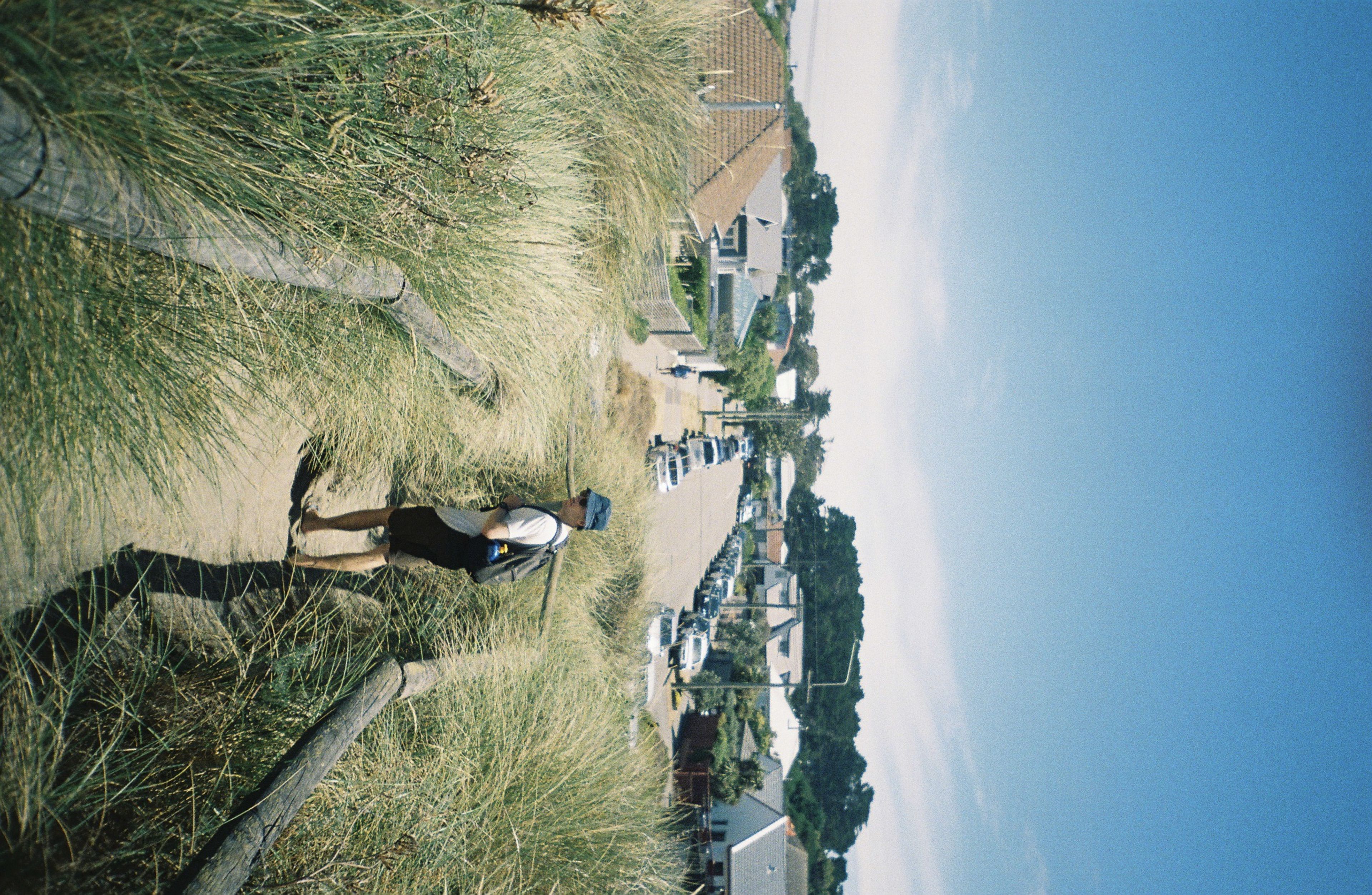 path grass person hat backpack houses road cars sky blue trees sand fence walkway summer outdoor nature landscape