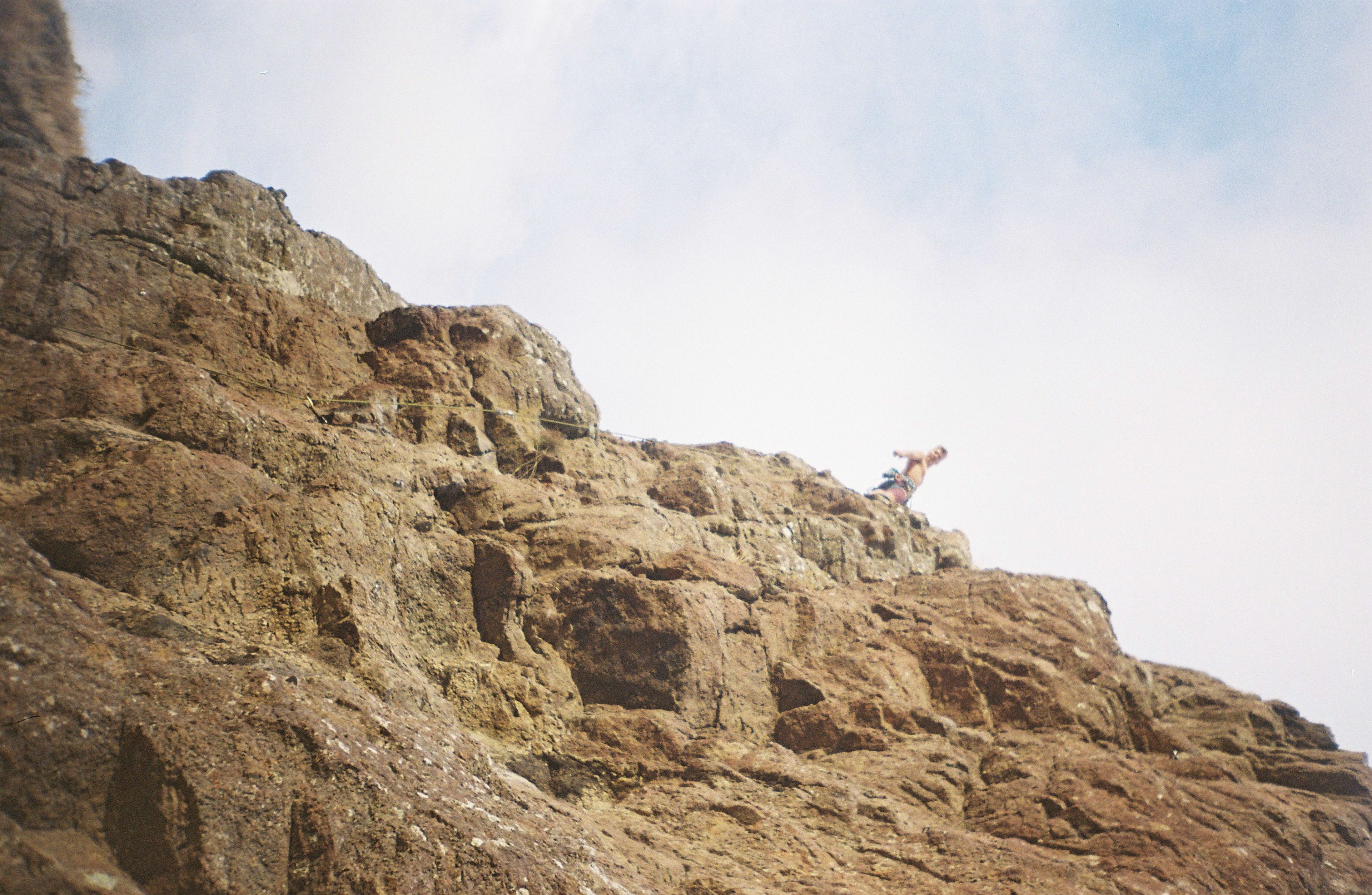 rock cliff climbing person sky clouds brown blue rugged outdoor adventure nature vertical height landscape formation texture ruggedness edge summit rocky surface elevation exploration challenge ascent precipice terrain geological feature cliffside craggy steep ruggedness outdoorsman daring verticality