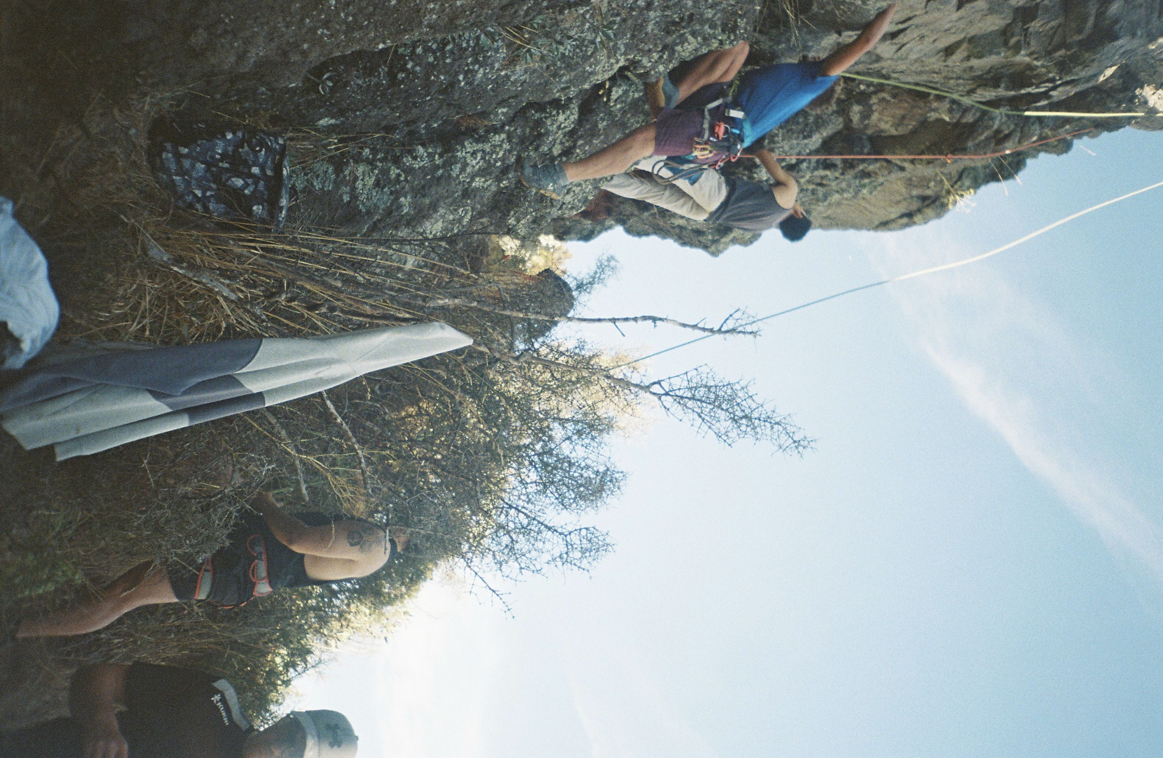 Climbing Rock Climber Rope Harness Helmet Outdoor Nature Sky Blue Cliff Vegetation Bushes Gear Equipment Adventure Sport Activity Challenge Vertical Ascent Safety Protection Teamwork Group Effort Exercise Fitness Rugged Terrain Exploration Courage Determination