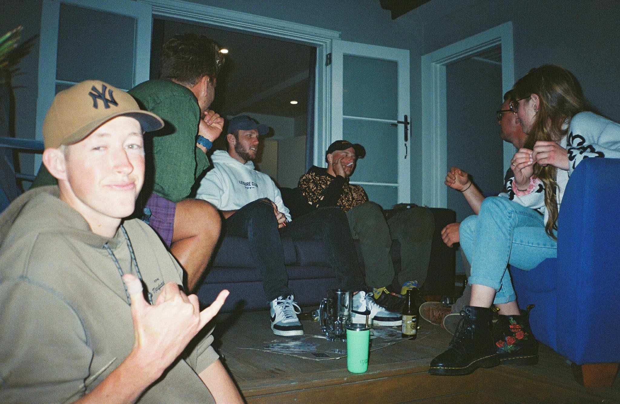 people group hats caps couch table drinks bottles glasses window door blue brown green black white sneakers jeans jacket shirt shorts sneakers boots lamp plant rug casual indoor gathering