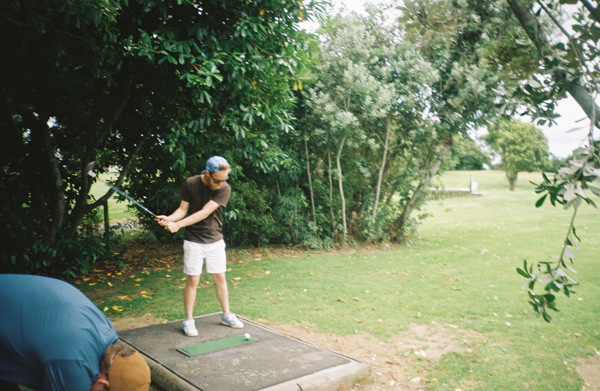 golf player swing club trees grass outdoors green shorts cap shirt shoes tee nature foliage bushes leaves pathway standing action summer sports recreation leisure activity