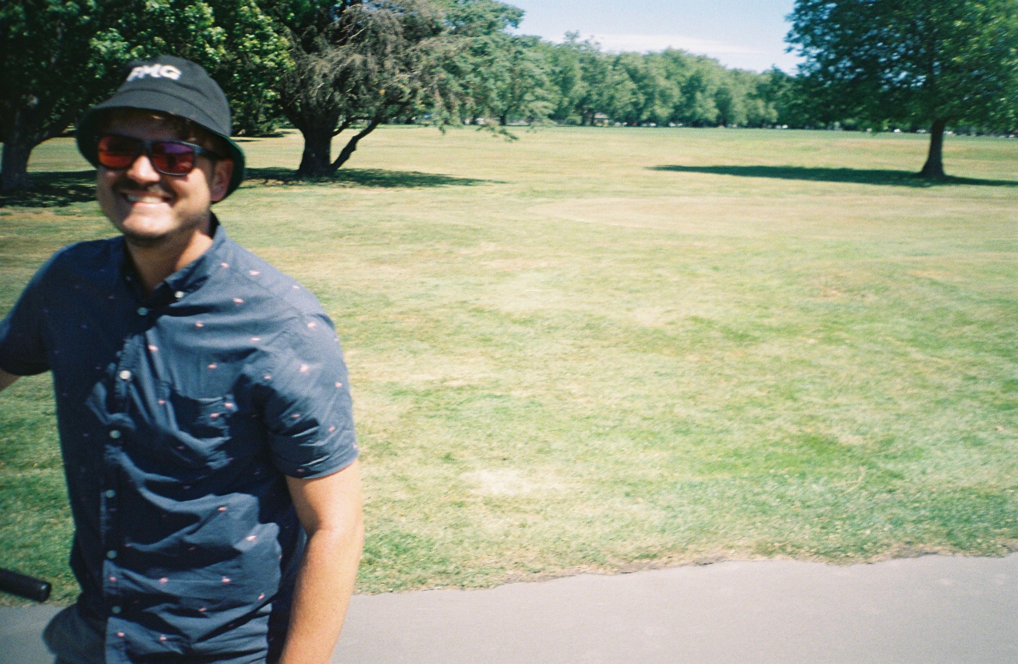 man hat sunglasses shirt grass trees park outdoors summer green blue sky pathway shadow smile casual nature
