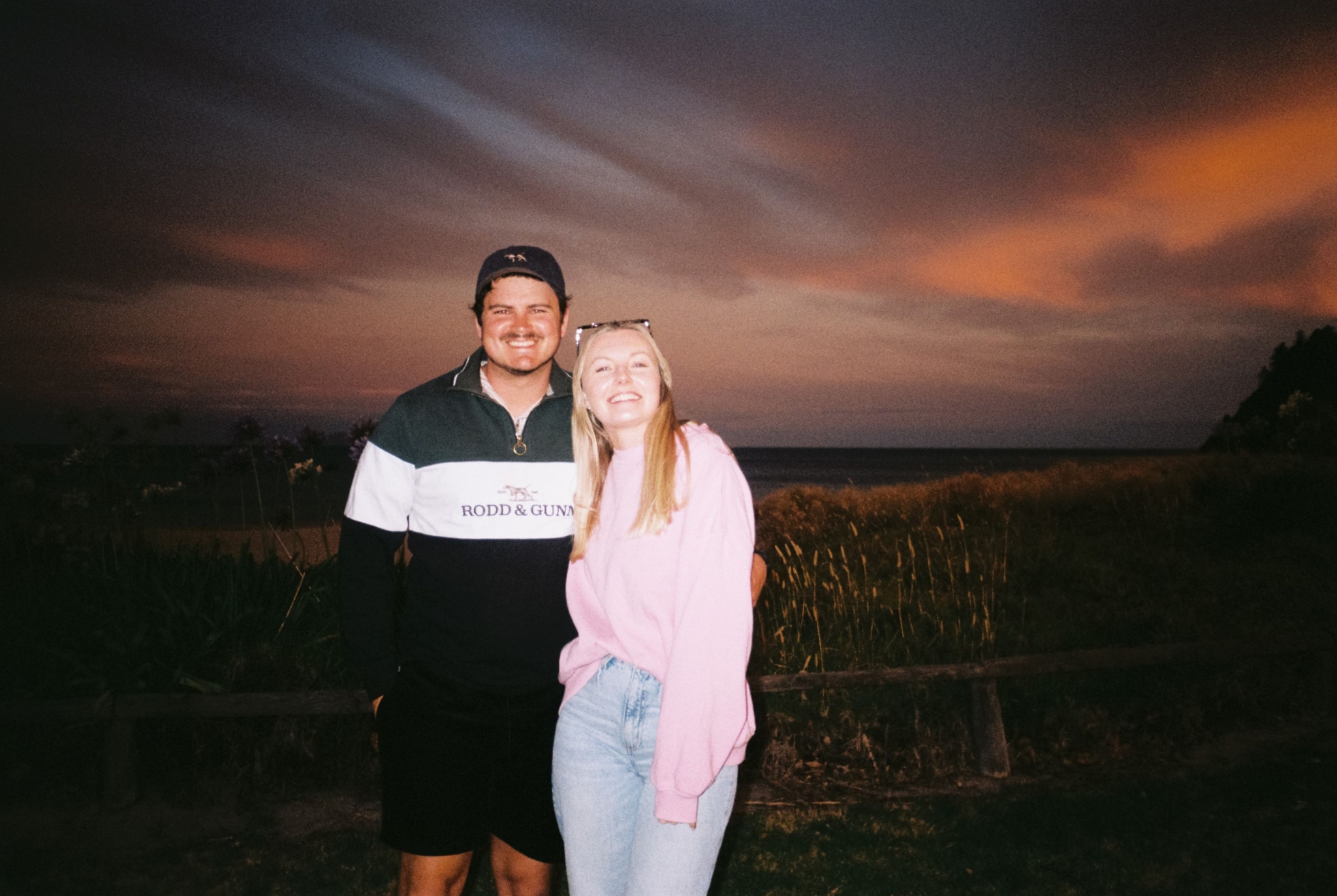 people couple sunset sky clouds grass fence jacket jeans hat smile pink black blue orange outdoor landscape twilight dusk