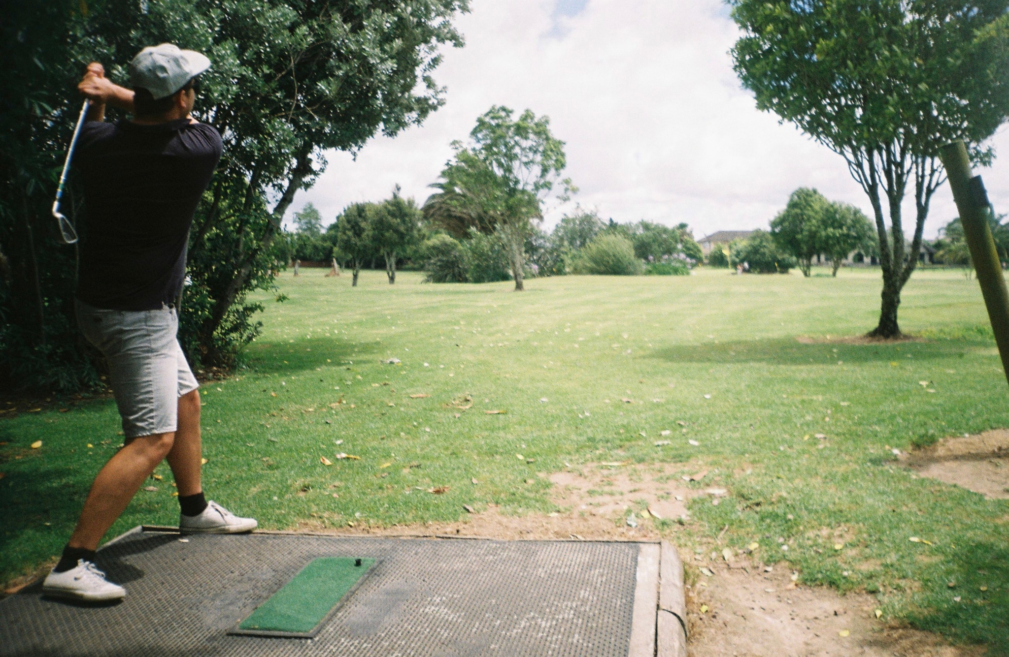 golfer golf swing club course grass trees sky outdoors sports green shorts shoes cap tee box summer fairway player action landscape recreation game activity nature leisure clouds