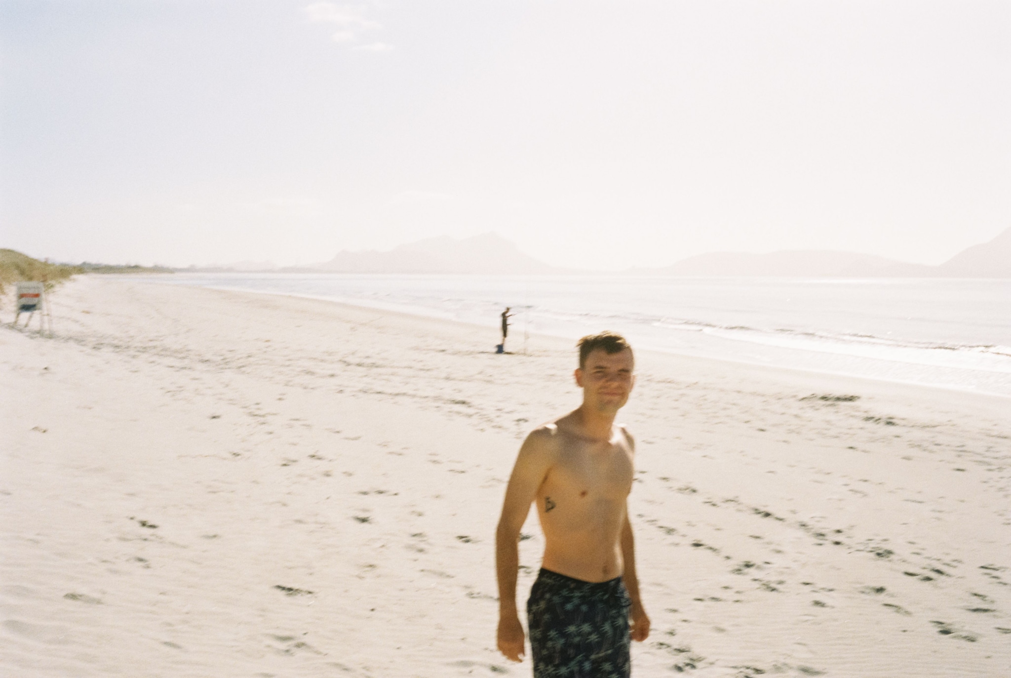 Beach Sand Ocean Water Sky Person Shirtless Shorts Sunlight Horizon Waves Footprints Distant Hills Coastline Shoreline Summer Outdoors Relaxation Leisure Scenic Tranquil Calm Bright Daytime Blue