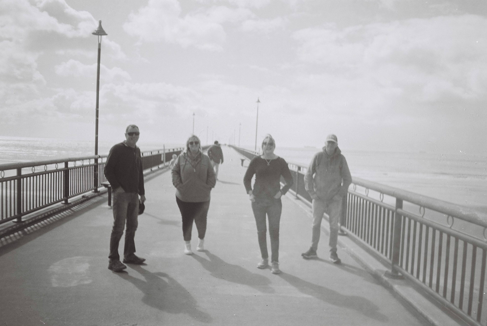 people bridge railing sky clouds lamps walkway black white grayscale shadows ocean horizon pathway