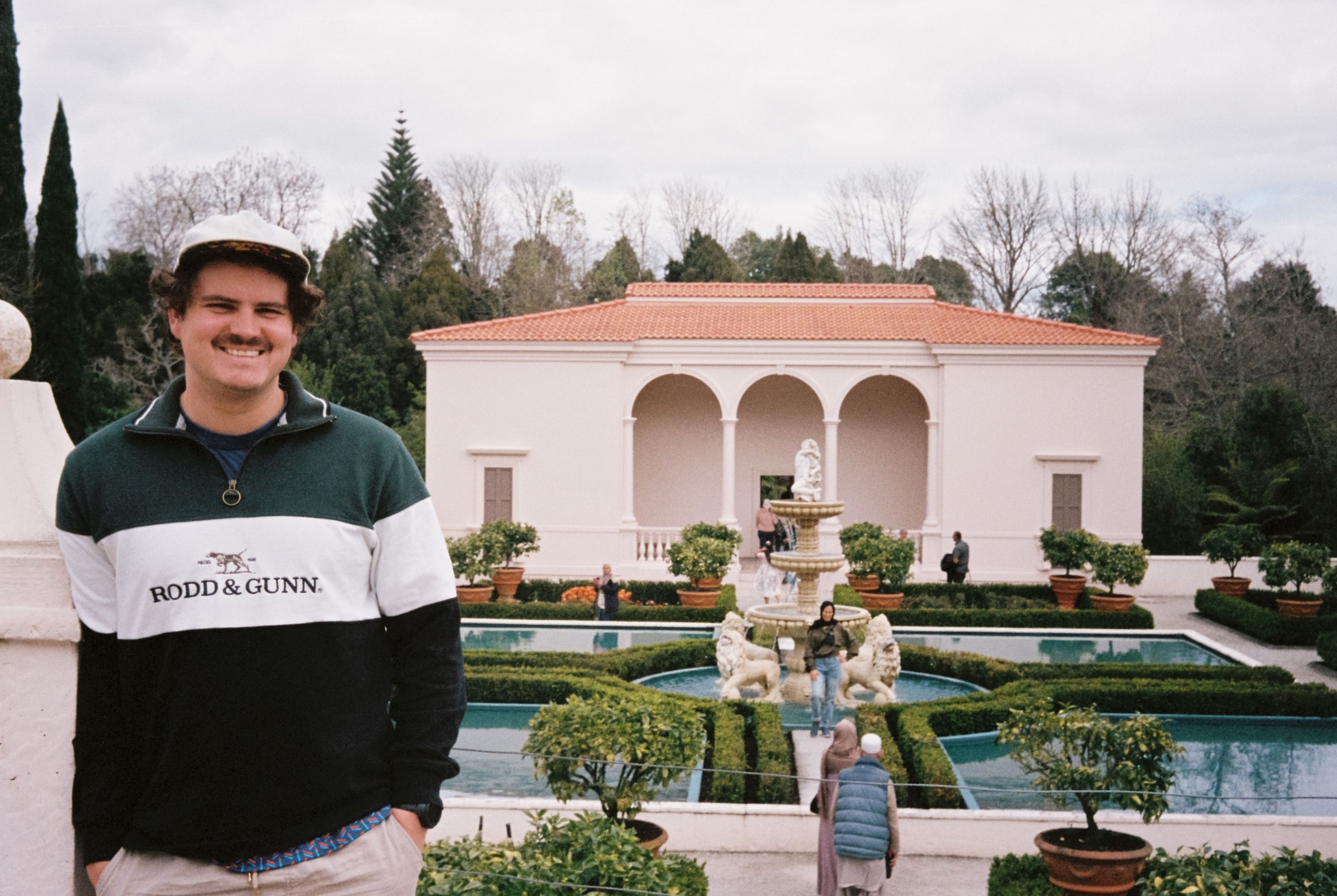 Man Garden Building Fountain Water Trees Bushes Sky Clouds Pathway Columns Archway Hat Jacket Smile Green White Red Stone Grass Shrubs Architecture Outdoors Landscape Sculpture Walkway Balustrade