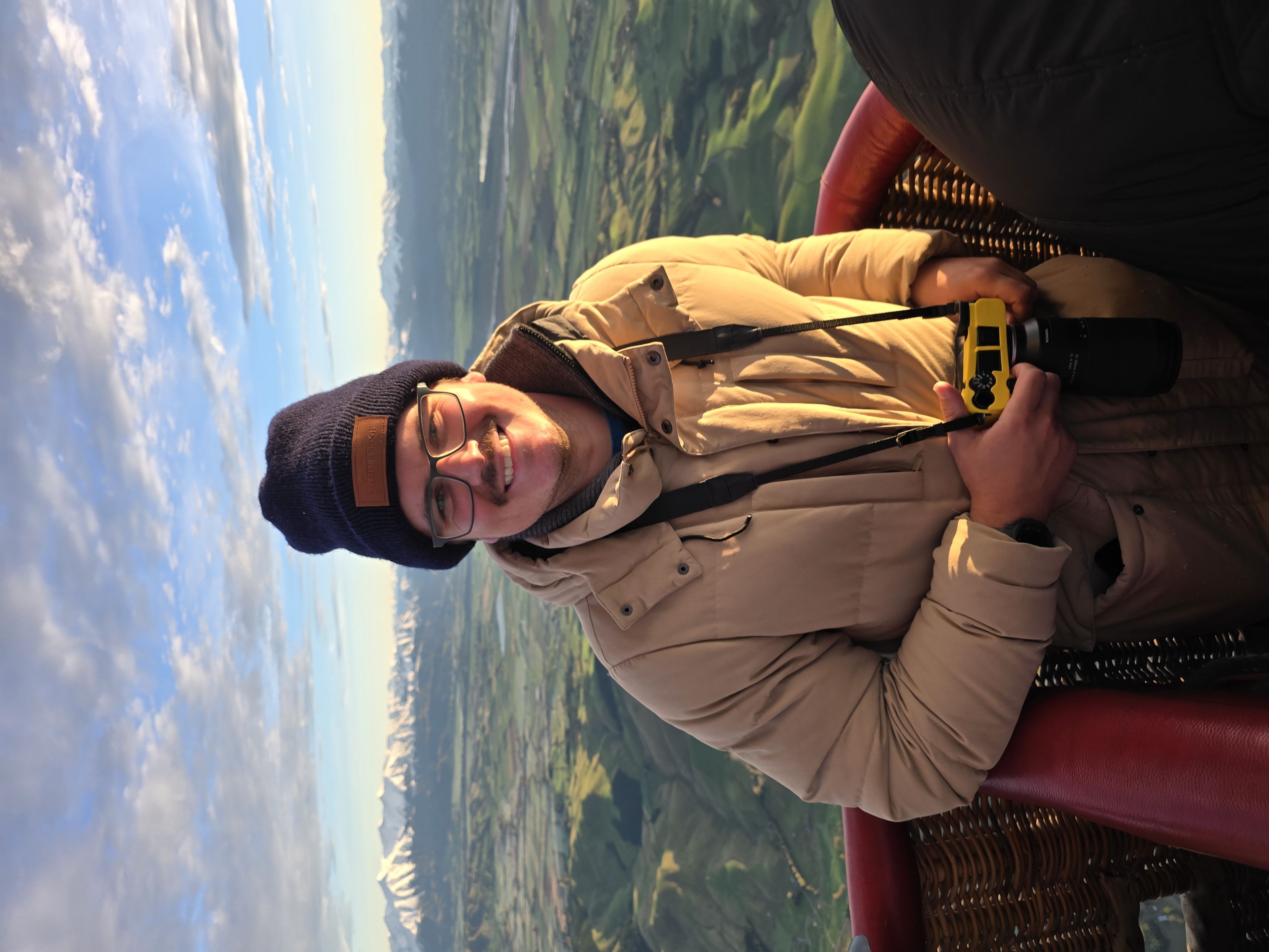 hot air balloon basket person coat beanie hat glasses camera landscape sky clouds mountains snow green fields horizon sunrise morning outdoor adventure travel photography smile red blue beige black yellow