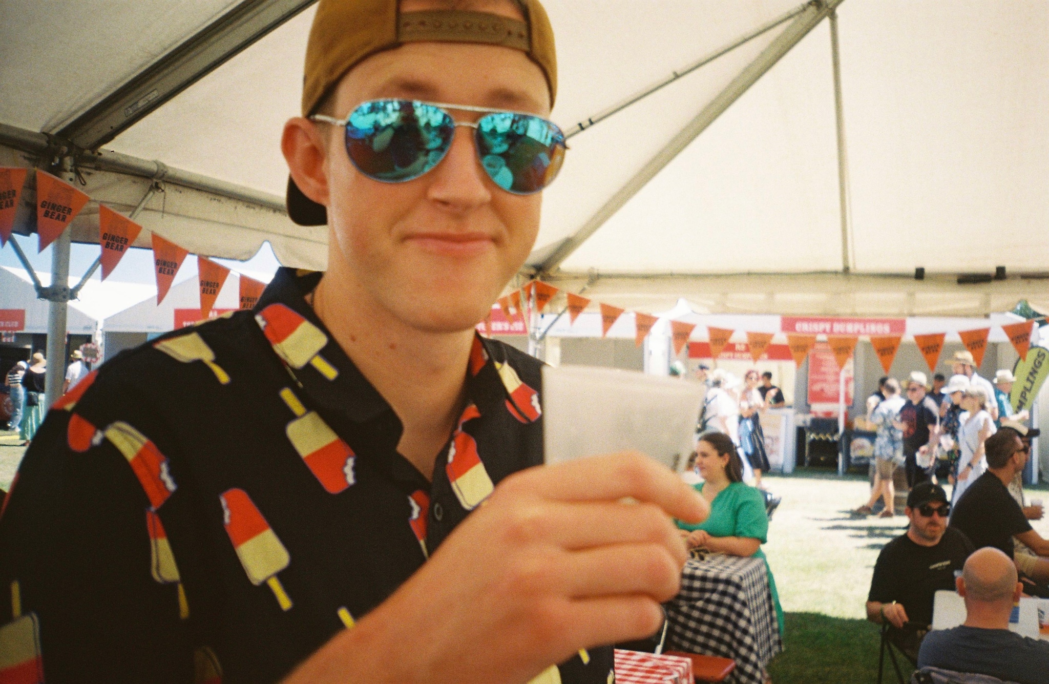 Man Hat Sunglasses Shirt Popsicle Drink Cup Tent Festival People Banner Table Grass Outdoors Daylight Smiling Casual Summer Event Celebration Gathering Relaxation Fun Leisure Enjoyment Socializing Fashion Style Accessories Primary Color: Blue