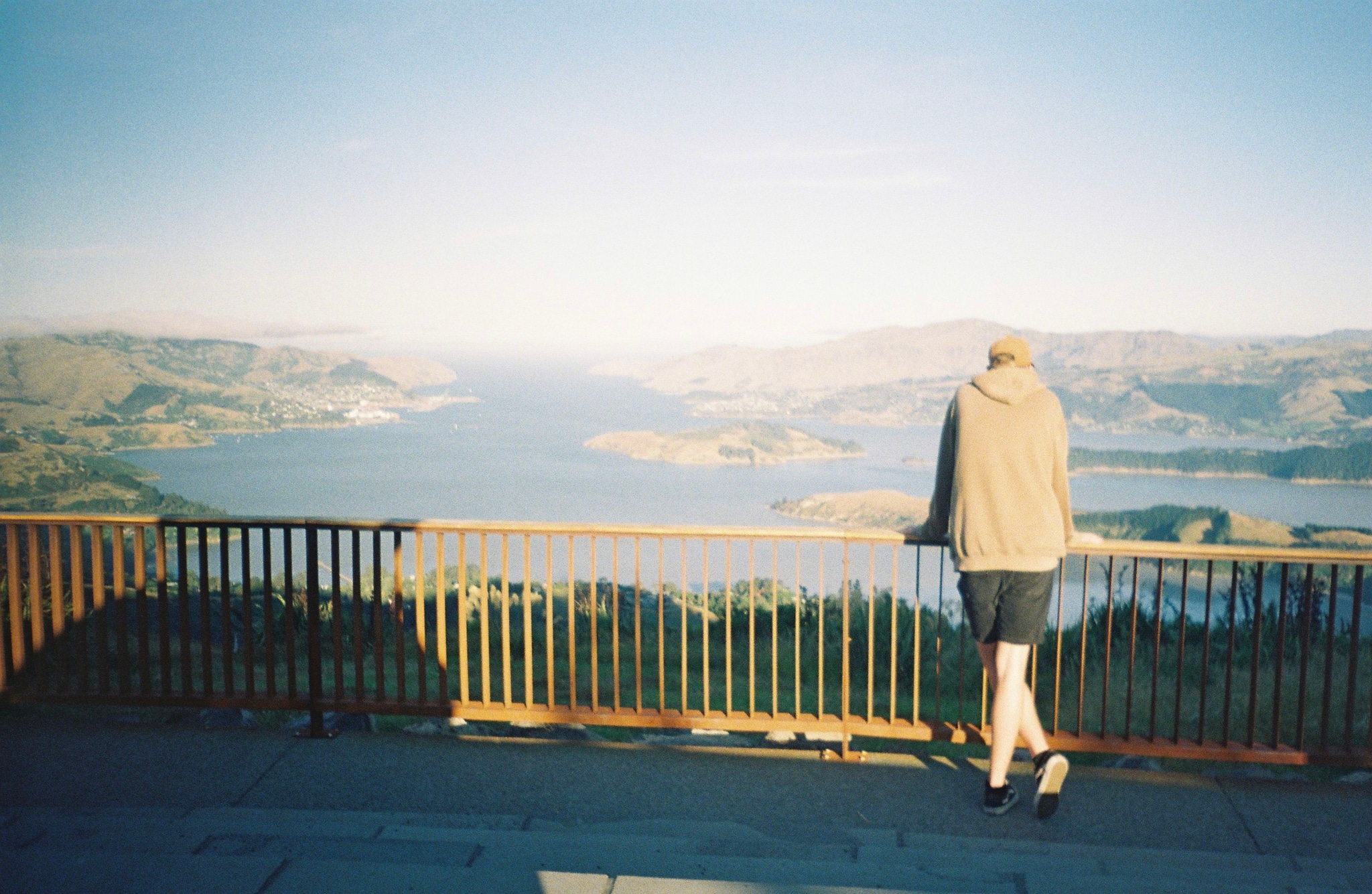 Person Landscape Viewpoint Railing Water Lake Mountains Hills Sky Horizon Blue Green Brown Hoodie Shorts Shoes Standing Balcony Nature Scenic Peaceful Calm Outdoors Daylight