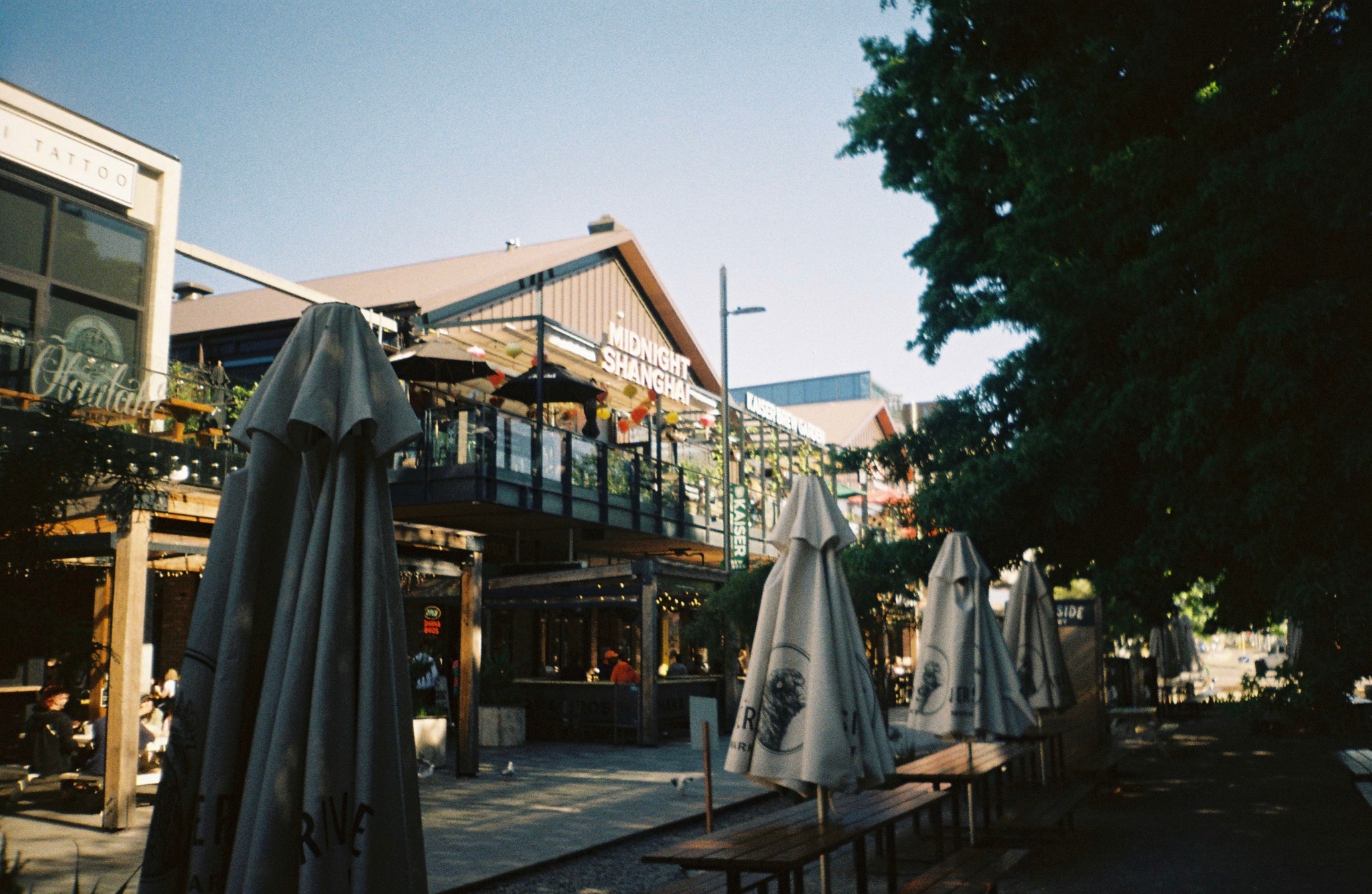 outdoor umbrellas tables chairs building balcony windows trees sky sunlight shadows street architecture railing plants blue
