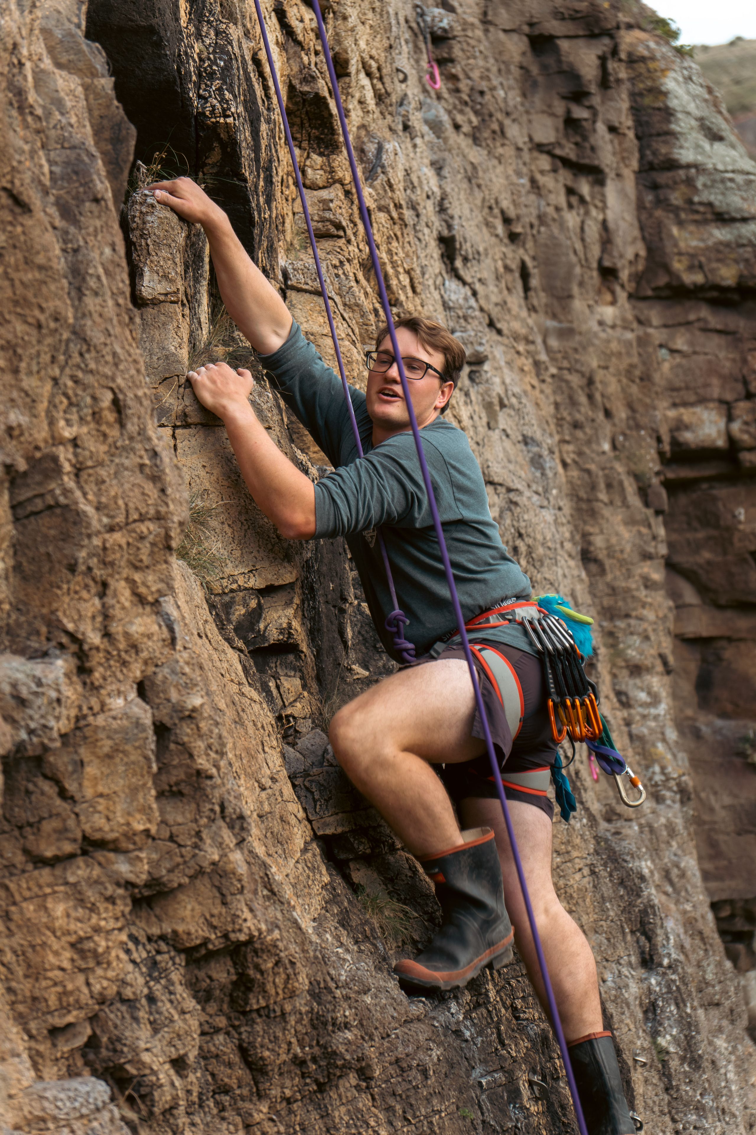rock climbing climber rope harness helmet rock wall cliff outdoor adventure sport safety gear boots brown green blue gray rugged ascent challenge vertical nature equipment protection concentration effort determination focus strength balance technique exploration physical activity recreation primary color brown