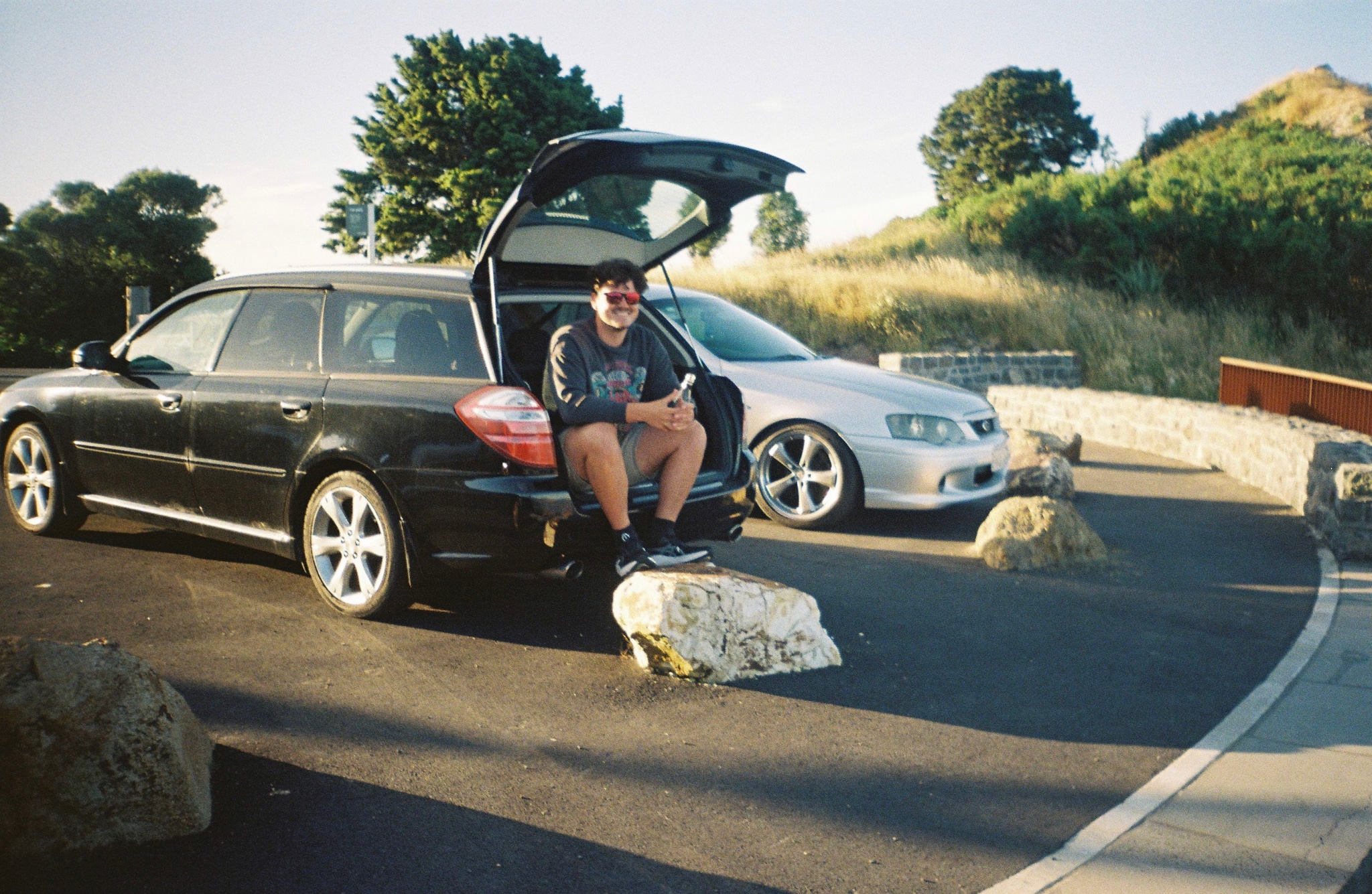 car vehicle person sitting road rock tree grass sky outdoor black white silver blue green stone hill landscape parking sunlight shadow casual relaxed summer travel journey transportation nature scenery