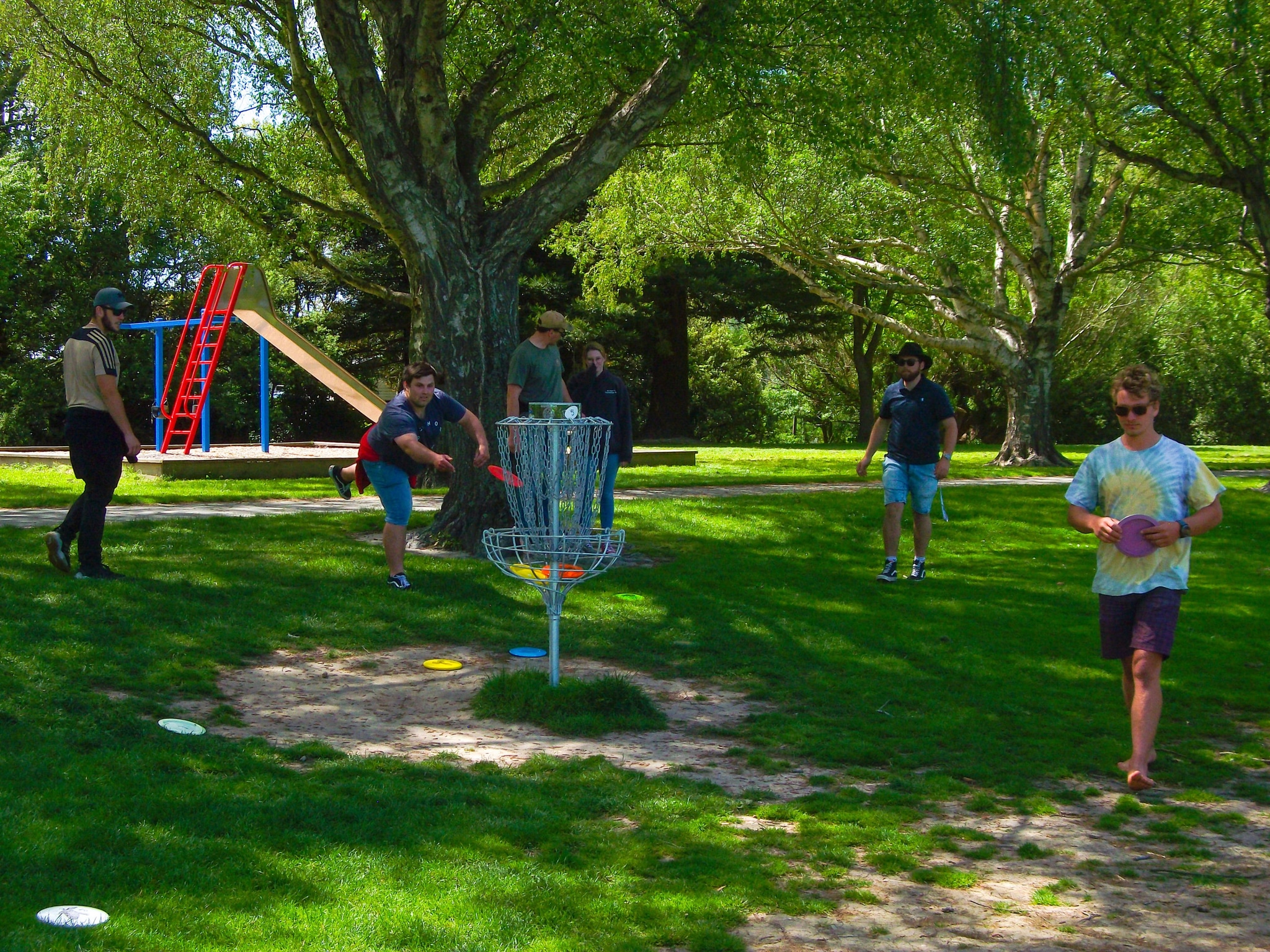 park trees grass people disc golf basket frisbee playground slide sunlight shadow green red blue brown black white casual clothing outdoors recreation leisure activity